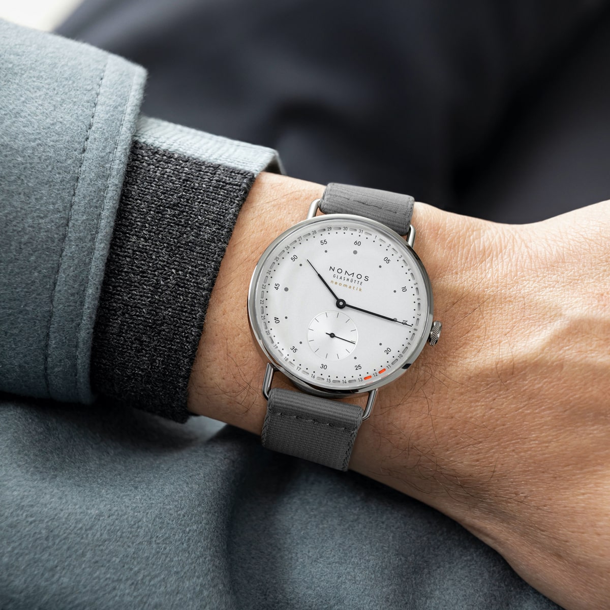 Eine Person trägt eine stilvolle NOMOS Metro-Armbanduhr mit minimalistischem Design, grauem Armband und silbernem Gehäuse. Die Uhr hat ein weißes Zifferblatt, ein kleines Sekunden-Hilfszifferblatt und feine schwarze Stundenmarkierungen, die perfekt zum hellgrauen Stoff des Ärmels der Person passen.