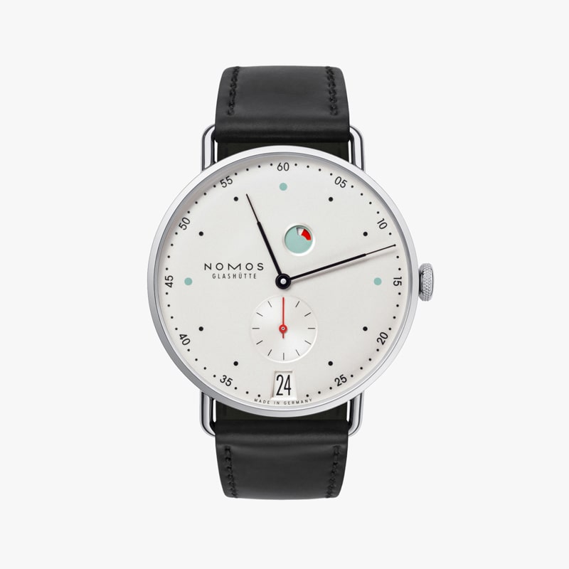 Die NOMOS Metro ist eine minimalistische Armbanduhr mit weißem Zifferblatt, Datumsfenster und kleinem Sekundenzeiger. Sie verfügt über eine markante rote und blaue Anzeige oben, eingebettet in ein poliertes Silbergehäuse mit dünnen schwarzen Stundenmarkierungen auf einem eleganten schwarzen Lederarmband.