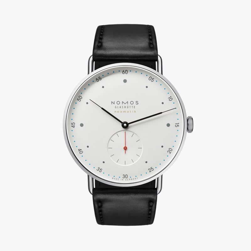 Die NOMOS Metro ist eine minimalistische Armbanduhr mit weißem Zifferblatt, schwarzem Lederarmband und silbernem Gehäuse. Sie verfügt über ein kleines Sekunden-Hilfszifferblatt bei 6 Uhr, dünne Zeiger und dezente Stundenmarkierungen. Die Marke „Nomos Glashütte“ ist elegant auf dem Zifferblatt abgebildet.