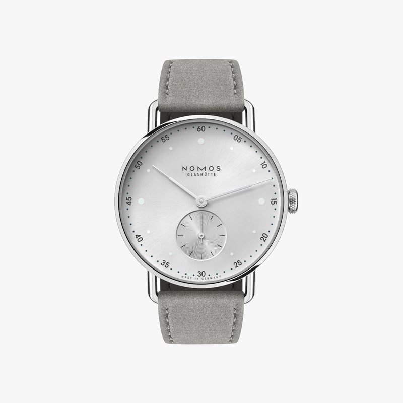 Die NOMOS Metro ist eine elegante, minimalistische Armbanduhr mit silbernem Zifferblatt und grauem Lederarmband. Sie verfügt über einfache Stundenmarkierungen, ein kleines Hilfszifferblatt für die Sekunden und dünne schwarze Zeiger. Der Markenname „NOMOS Glashütte“ ist elegant auf das Zifferblatt gedruckt.