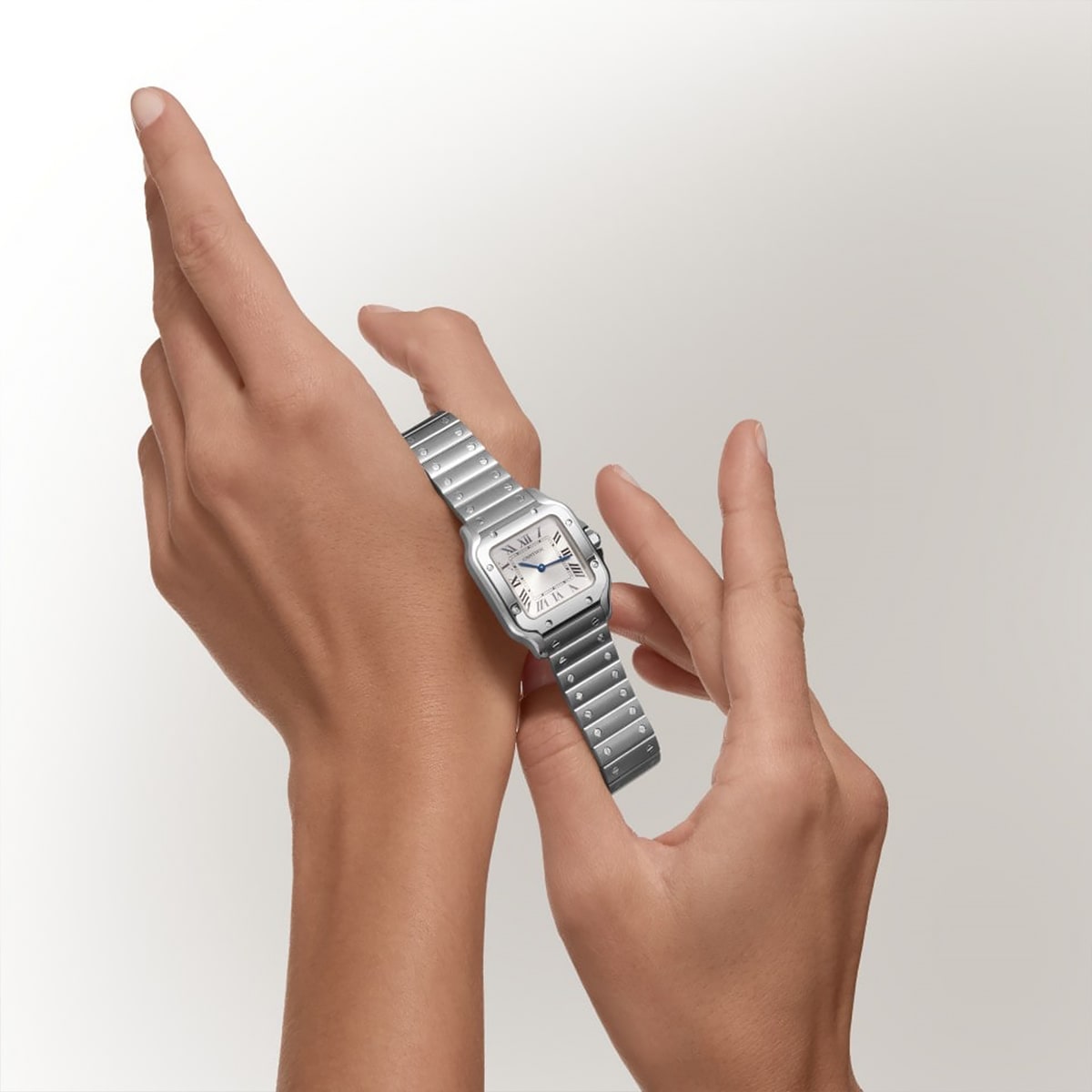 Die linke Hand einer Person trägt eine silberne Metallarmbanduhr der Santos Kollektion mit quadratischem Zifferblatt und römischen Ziffern, während die rechte Hand die Uhr einstellt. Der Hintergrund ist einfarbig und hell.