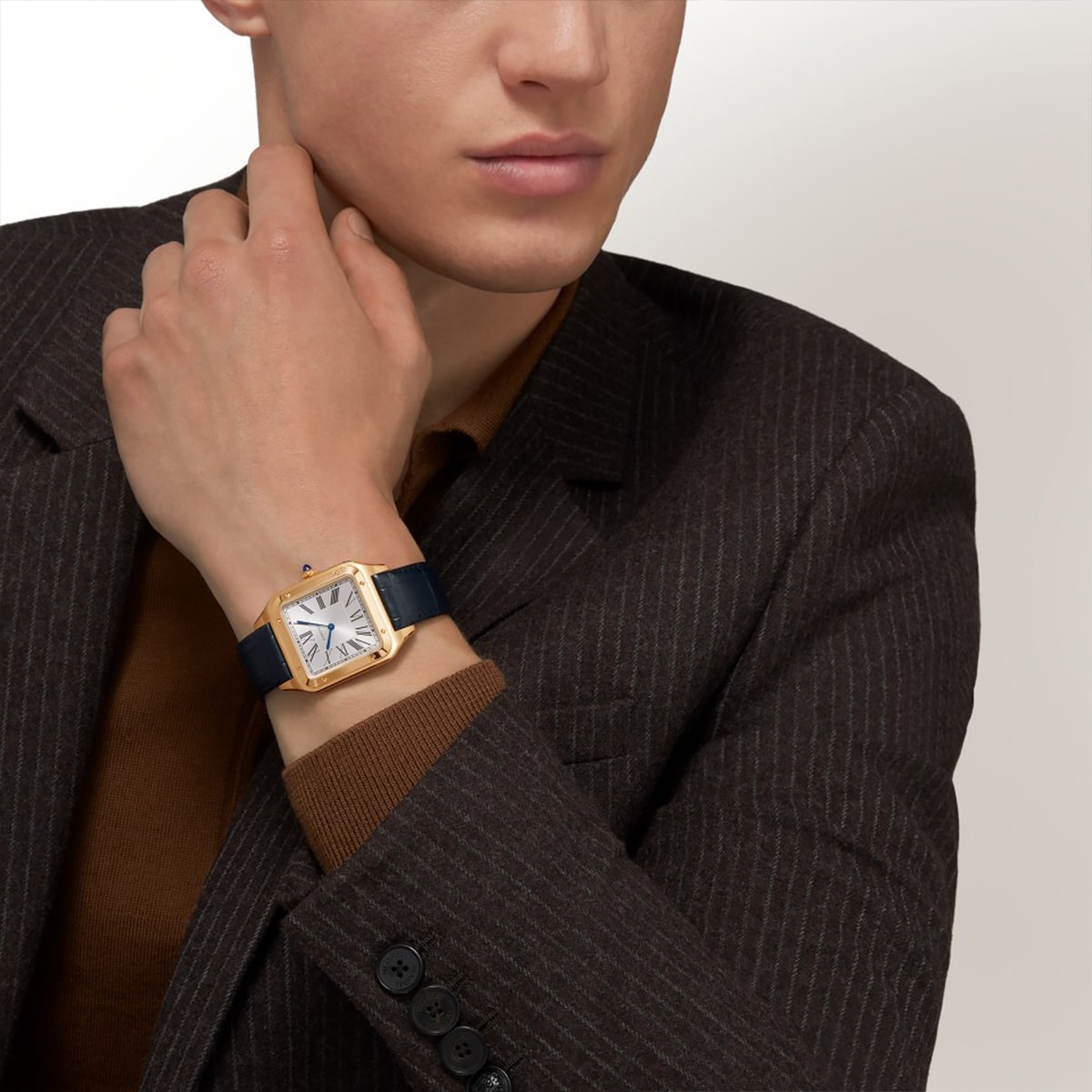 Ein Mann, der einen braunen Nadelstreifenanzug und ein braunes Hemd trägt, berührt seinen Hals und präsentiert eine goldene Armbanduhr der Santos Kollektion mit quadratischem weißem Zifferblatt und schwarzem Lederarmband an seinem linken Handgelenk.