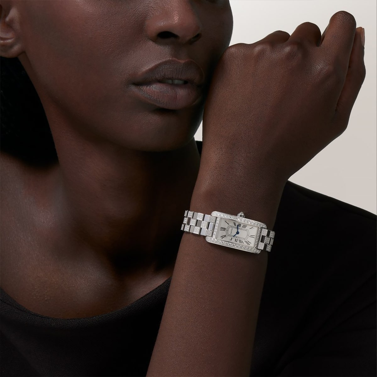 Eine Person mit dunkler Hautfarbe, die ein schwarzes Oberteil trägt, zeigt eine silberne rechteckige Armbanduhr der Tank-Kollektion mit einem Metallband an ihrem linken Handgelenk und stützt ihr Kinn auf ihre Hand vor einem hellen Hintergrund.