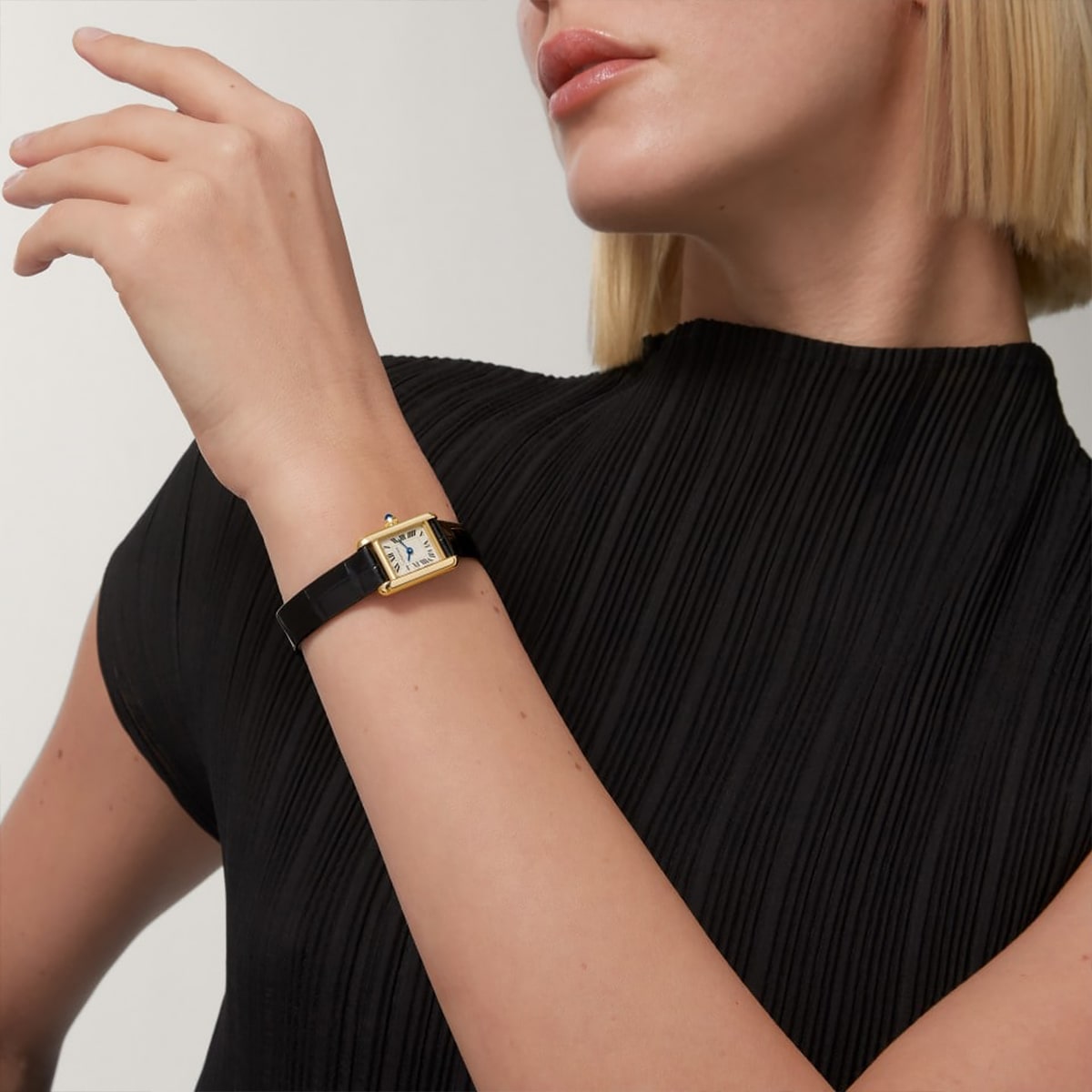 Eine Frau in einem schwarzen, in Falten gelegten Oberteil trägt eine goldene Uhr mit quadratischem Zifferblatt und schwarzem Lederarmband von Tank Kollektion an ihrem linken Handgelenk. Ihr blondes Haar ist zu einem stumpfen Bob geschnitten, wobei ihr Gesicht teilweise sichtbar ist.