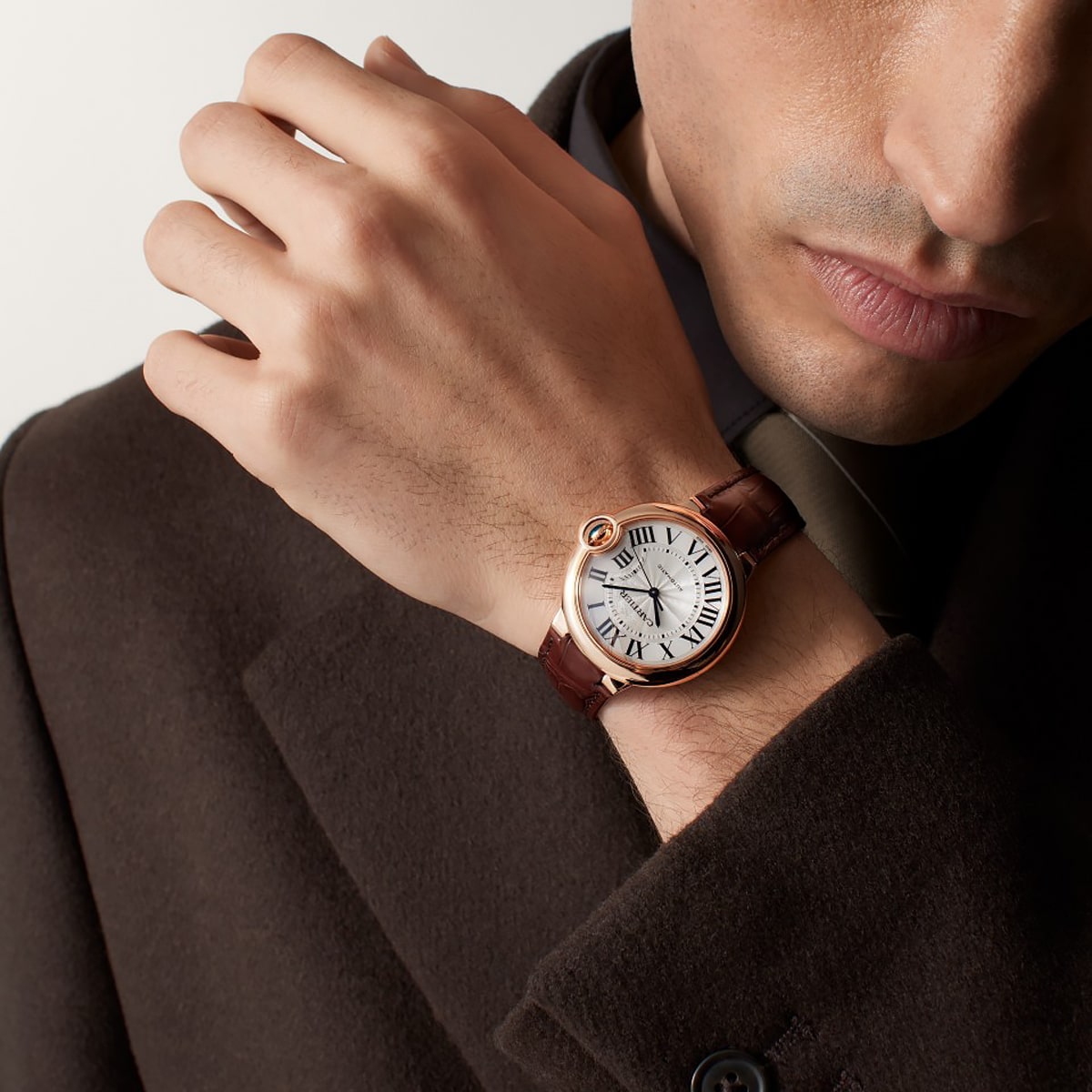 Ein Mann im braunen Anzug und mit Krawatte trägt eine Cartier-Luxusarmbanduhr mit rundem, silbernem Zifferblatt, römischen Ziffern und einem braunen Lederarmband an seinem linken Handgelenk.
